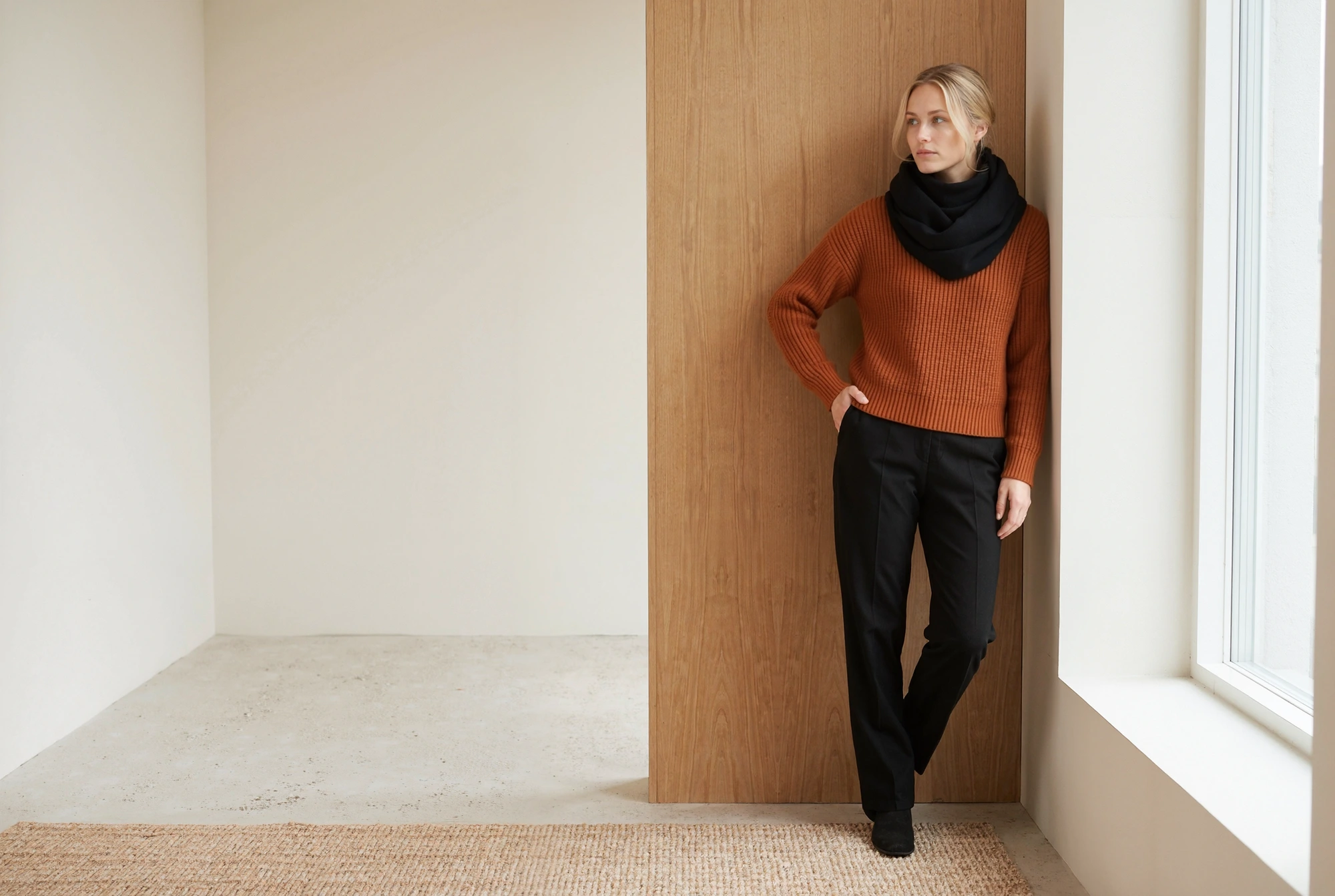 Model wearing a black knit scarf and a rust sweater, leaning against a wood panel.